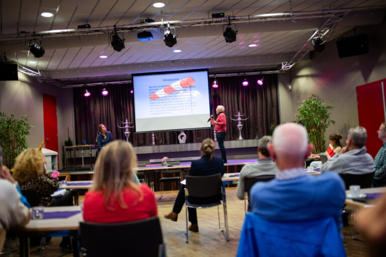 Foto van achter uit de zaal kijkend naar het podium waar iemand een presentatie houdt. Op de voorgrond zitten mensen te kijken naar de presentatie.